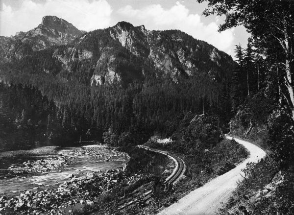 A view of Mount Persis, part of the Cascade Range in Washington. Railroad tracks are running along a low and rocky river, and along the tracks runs a dirt road winding into the distance.