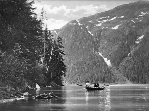 View across water toward three men in a rowboat on a river in a mountain gorge.