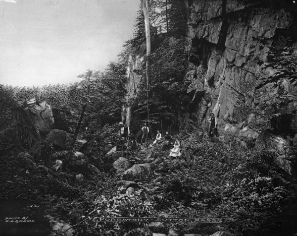 Three men and five women posing in a rocky, wooded ravine. Caption reads: "Purgatory- Sutton, Mass."