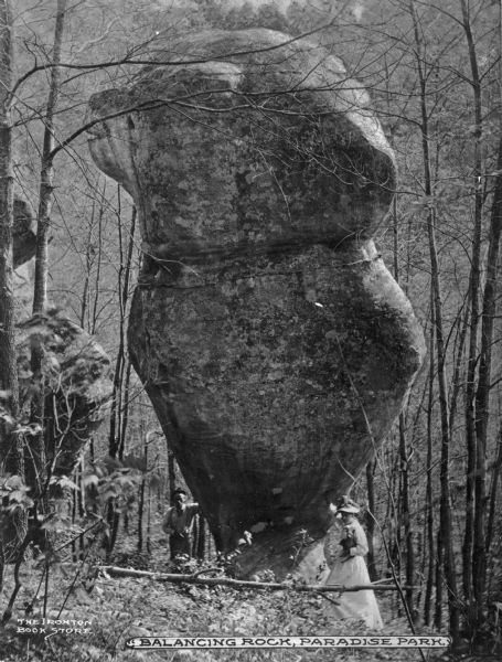 A large, balancing boulder in Paradise Park, Ohio. A man is posed standing on the left side of the rock and a woman is posed standing on the right side of the rock holding a small dog in her arms. Caption reads: "Balancing Rock, Paradise Park."
