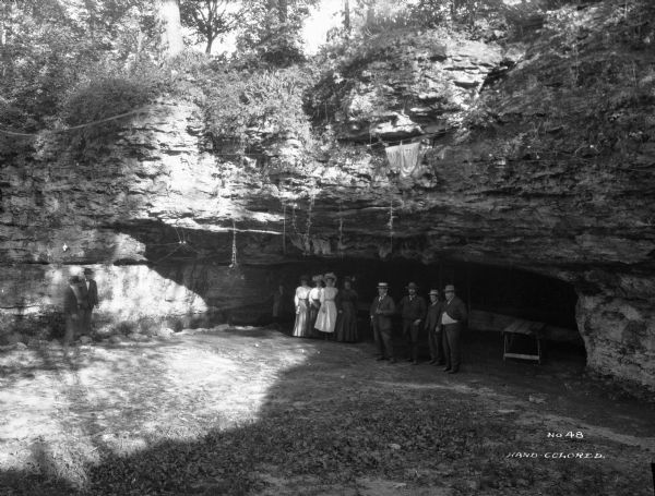 View of six men, four women, and a boy standing in front of an entrance to a cave in Doling Park.