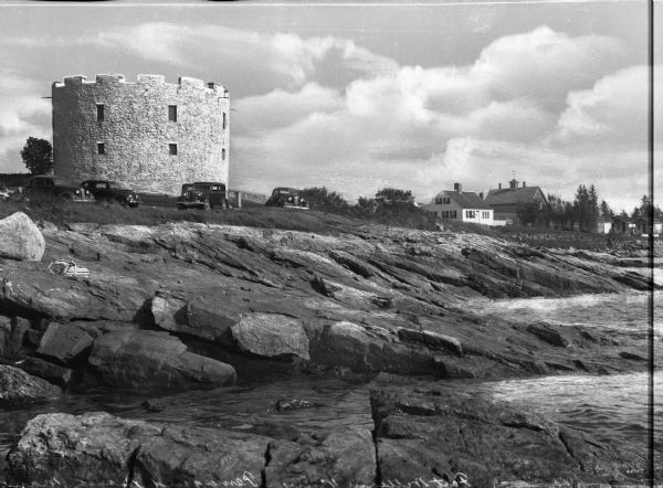 View from rocky shore toward the stone tower at Fort William Henry. Four automobiles are parked in front of the tower, with houses and other buildings in the background on the right.