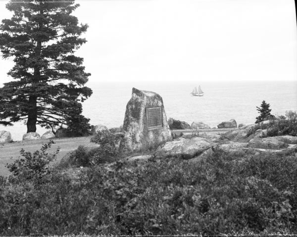 View across shrubs toward a stone historical marker dedicated to Champlain, a Canadian explorer. The marker overlooks the ocean, and a sailboat is visible in the background.