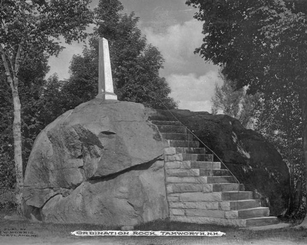 A view of Ordination Rock, with an obelisk sitting on top of a flat topped erratic boulder with stairs up the side of it. This is where on September 12th, 1792, Rev. Samuel Hidden was ordained as a pastor. Caption reads: "Ordination Rock, Tamworth, N.H."