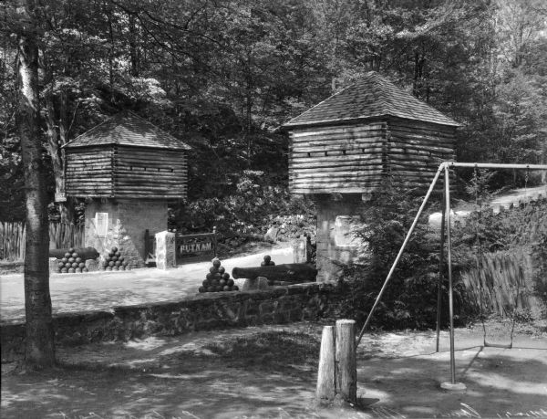 A view of the main entrance to Putnam Park, including two wooden towers on either side of the road, various cannons and cannon balls. There is a swing set in the right foreground.