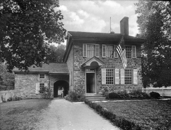 A view of Washington's headquarters in Valley Forge, 1777-1778, a stone house with a large colonial flag displayed from the front.