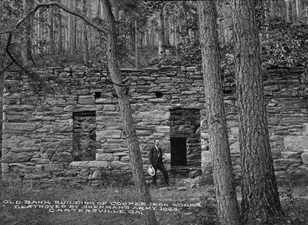 A man standing in front of the old stone bank building of Cooper Iron Works. Caption reads: "Old Bank Building of Cooper Iron Works, Destroyed by Sherman's Army, 1864, Cartersville, GA."