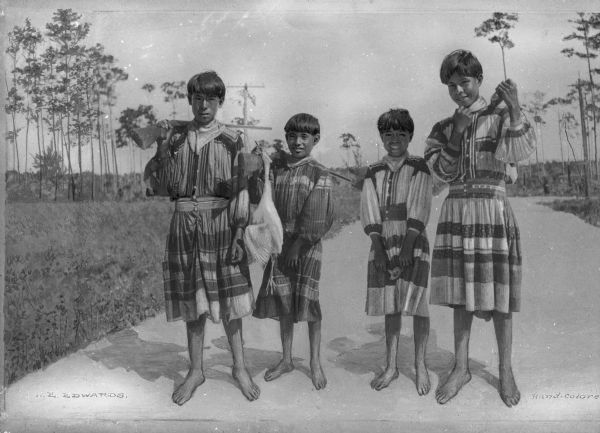 A group of four Seminole Native American youth standing on a path with trees behind them. Three of them are holding rifles and one is holding a dead bird.