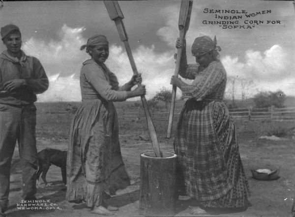 A view of two Seminole Native American women grinding corn for "Sofka," a meat stew thickened with vegetables and meal. A man and dog are on the left. Caption reads: "Seminole Indian Women Grinding Corn for 'Sofka'."
