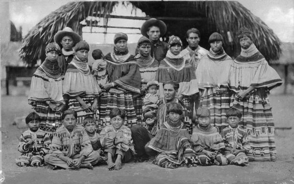 A group of Seminole Native American men, women, and children in front of a structure with a thatched roof in the Everglades area. There are twenty individuals in total, and all are wearing traditional clothing and some are wearing jewelry.