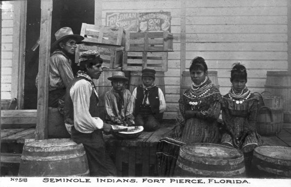 A group of Seminole Native Americans consisting of two men, two boys and two girls sitting outside on the porch of a building with one man cutting what looks to be a watermelon. Also on the porch are some crates and barrels and two signs advertising "Friedman Shoes" and "Hamilton-Brown's 2.50 Shops." Caption reads: "Seminole Indians, Fort Pierce, Florida."