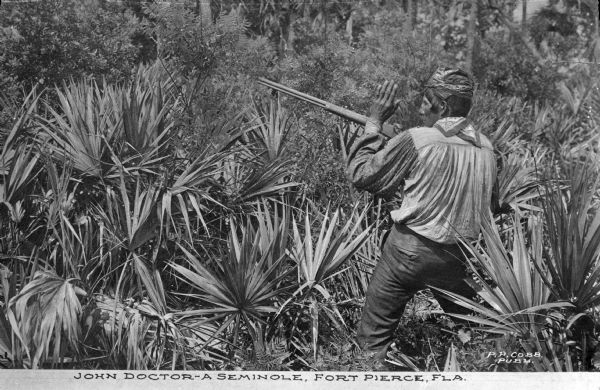 View of a Seminole Native American, John Doctor, holding a gun and standing in an area dense with plants. Caption reads: "John Doctor -- A Seminole, Fort Pierce, FLA."