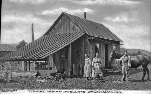 A view of a small wooden building with a group of people standing in front. A man on the right stands with a horse, and in the center are a woman, a young girl and a dog. Caption reads: "Typical Indian Dwelling -- Salamanca, N.Y."