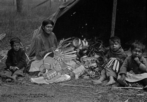A Native American woman weaving baskets outdoors. Three children are sitting nearby. Caption reads: "Indian Adepts, Glenn, Mich."