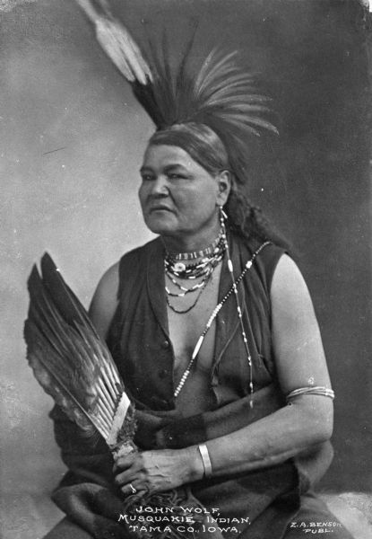 A portrait of John Wolf, a Musquakie Native American, wearing jewelry, a headdress, and holding a feathered object in his hands. Caption reads: "John Wolf, Musquakie Indian, Tama Co., Iowa."