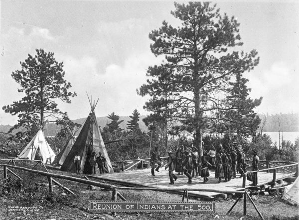 A view of people standing near three tepees, and with many Native Americans dancing in a circle on a wooden platform. Caption reads: "Reunion of Indians at the Soo."