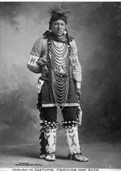 Full-length portrait in front of a painted backdrop of an Indian man in elaborate tribal costume in Alberta, Canada. Caption reads: "Indian in Costume, Medicine Hat, Alta."
