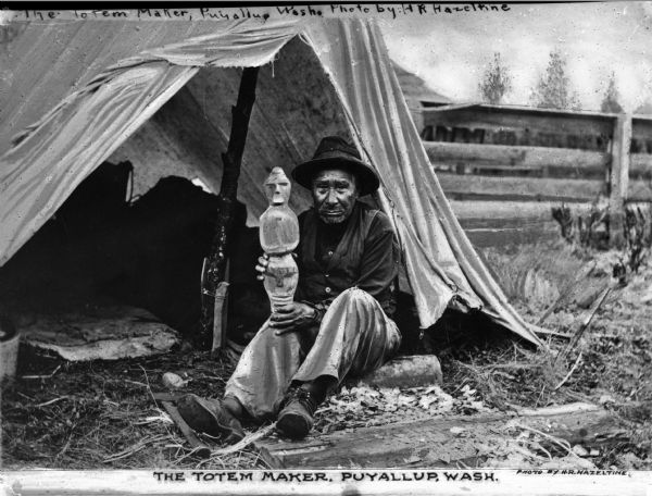 A view of a Native American man, the Totem maker, sitting outside of a tent and holding a carved totemic figure. Caption at bottom reads: "The Totem Maker, Puyallup, Wash."