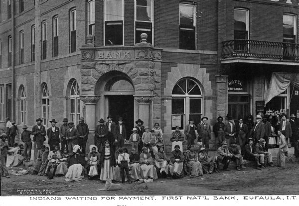 A group of Native American men, women, and children, waiting in front of the First National Bank for their payment, possibly in 1910. Caption reads: "Indians waiting for payment. First Nat'l Bank, Eufaula, I.T."