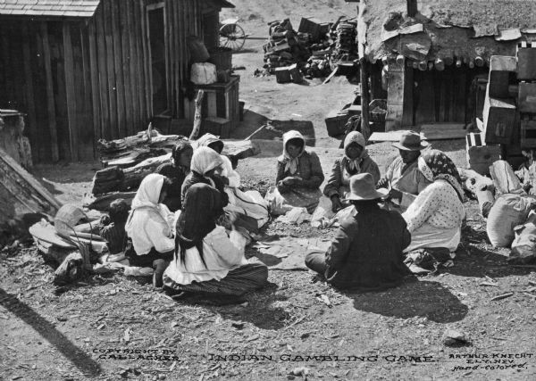 A group of Native Americans sitting in a circle outside of two structures while playing a gambling game. Caption reads: "Indian Gambling Game."