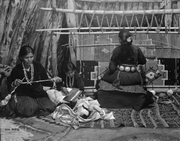 View of three Pueblo Native American women. The woman on the left is kneeling and spinning wool, and a young girl in the center is sitting and combing wool. The woman on the right is working on a loom.