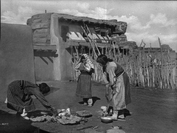 Three Pueblo Native American women, standing outside of an adobe structure and around a fire pit.
