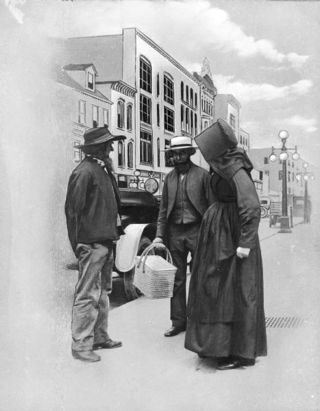 A view of two Amish men, one of whom is holding a basket, and an Amish woman standing on the sidewalk along a commercial street, with an automobile and streetlights in the background.