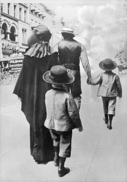 A view of an Amish man and woman with two children walking down the sidewalk along a street.
