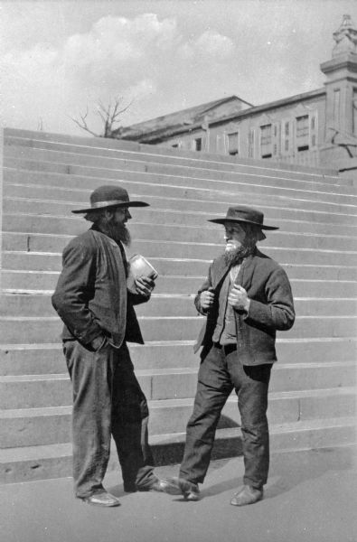 A view of two Amish men standing outdoors near the bottom of a flight of stairs. Buildings are in the background.