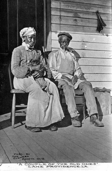 A portrait of an old African American couple sitting on a porch. The woman on the left is smoking a pipe and is holding a small dog in her lap, and the man is on the right is smoking a pipe. Caption reads: "'A Couple of the Old Ones' Lake Providence, LA."
