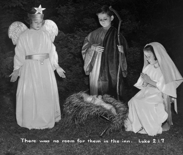 Children acting in a living nativity scene at St. Michael's Home. The three children represent an angel, Joseph, and Mary, and are gathered around a manger holding a doll. Caption reads, "There was no room for them in the inn. Luke 2:7."