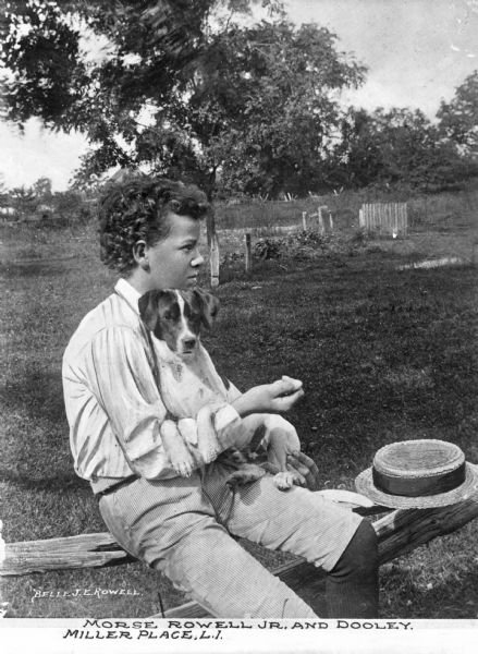 A portrait of Morse Rowell Jr. and a dog, Dooley. Morse is posed sitting on a fence and holding Dooley in his lap with a hat resting beside him. Caption reads: "Morse Rowell Jr. and Dooley. Miller Place, L.I."