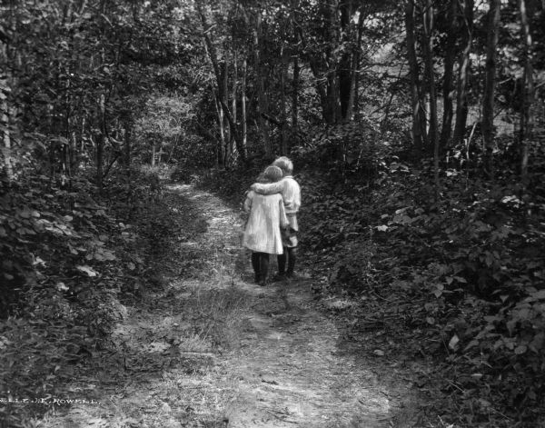 A view of a young girl and boy facing away and embracing on a path in dense woods.