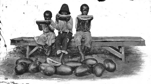 A view of three African American children seated on a long bench and eating watermelon, with a number of watermelons on the ground near their feet.