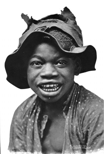 A studio portrait of a smiling African American boy wearing a tattered hat and an open shirt against a white background.
