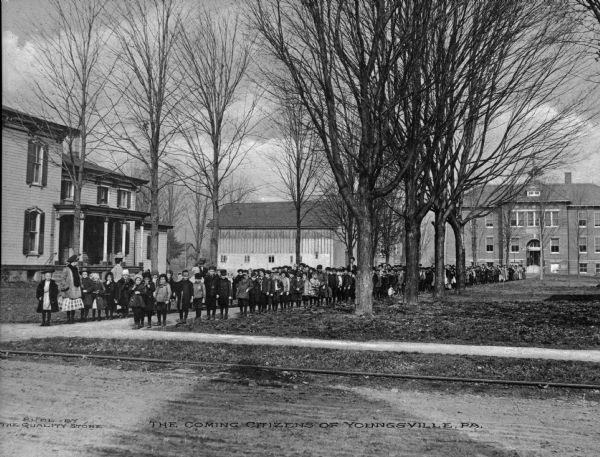 A view across road toward a group of school children lined up on a sidewalk. Caption reads: "The Coming Citizens of Youngsville, PA."