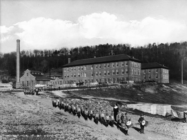 A view of St. Michael's Industrial School, with students lined up in formation along the road that leads to the school. Two boys with drums lead the procession.