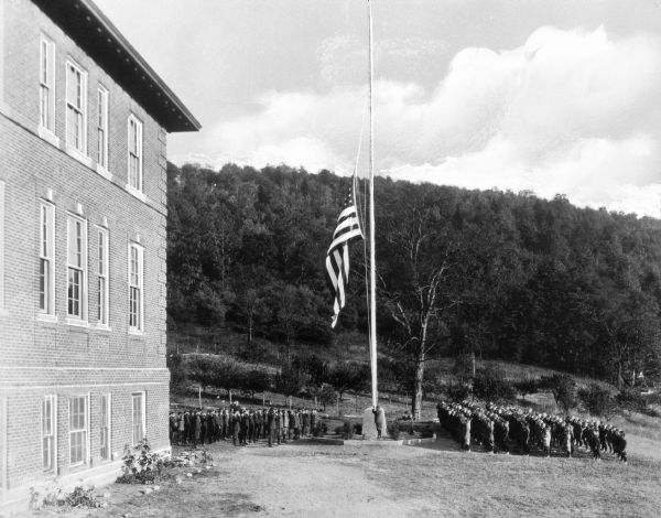 A view of the exterior of St. Michael's Industrial School, with students in formation saluting the flag being raised. There are trees on a hill behind them.