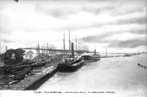 A view of boats laden with ore docked at the harbor. Caption reads: "The Harbor -- Looking Out, Lorain, Ohio."