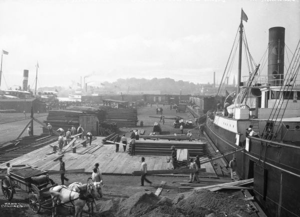 Boats unloading at Standard Oil Co.'s pier, with a horse, wagon, and a number of men handling lumber and other building materials.