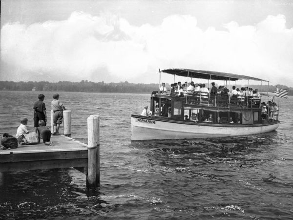 A view of an excursion boat, the "Dorianna," loaded with people on lake waters heading toward five young boys waiting on a dock.