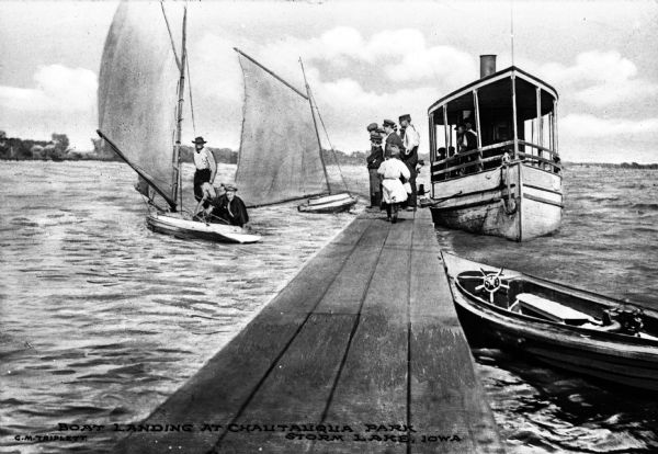 View down pier, with people standing at the end, and two sailboats in the water, with the one on the left manned by two men. There is an excursion boat docked at the end of the pier on the right. Caption reads: "Boat Landing at Chautauqua Park, Storm Lake, Iowa."