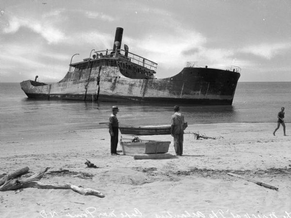 View from shoreline toward the wreck of the <i>S.S. Atlantus</i>, a concrete hulled ship in the water near a shoreline. In the foreground are three men on the beach near small boats.