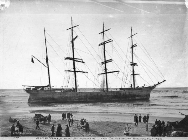 Elevated view across beach toward the ship beached in shallow water. There is a crowd of people on the beach in the foreground. Caption reads: "Ship 'Galena' Stranded on Clatsop Beach, Ore."