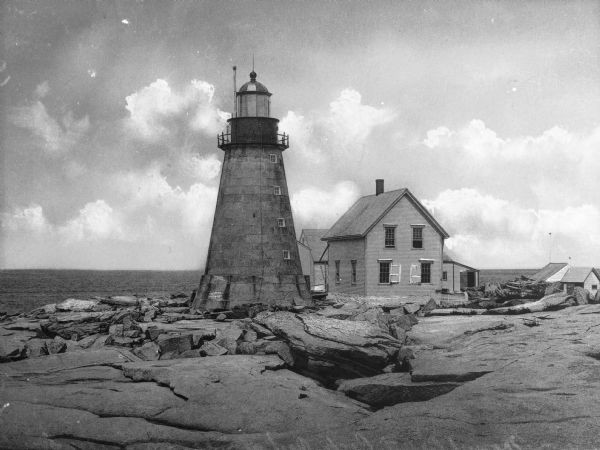View across rocks toward the Mount Desert Rock granite lighthouse, established in 1830 and automated in 1977, located thirty miles out to sea on a treeless island. The keeper's house and other outbuildings are just behind the lighthouse on the right.