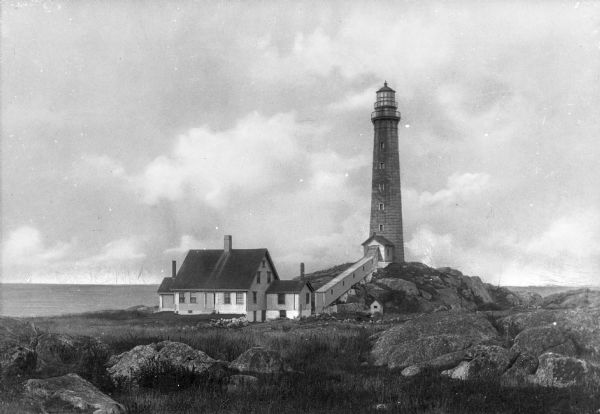 View across rocky ground and grass toward the Northern Lighthouse and the nearby cottage. A covered walkway leads from the house up to the entrance of the lighthouse.