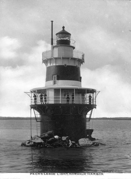 View across water toward six men standing on a deck with railings on Peck Ledge Lighthouse in Norwalk Harbor, put into operation in 1906. Caption reads: "Peck's Ledge Light, Norwalk Harbor."