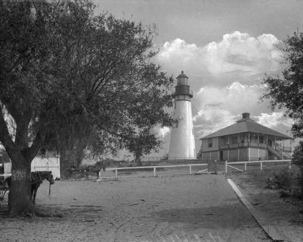 View along a sandy pathway toward the Amelia Island lighthouse near the keeper's house. Trees line the pathway, and two horses pulling a carriages are on the elft.

The lighthouse is constructed of brick – a “double-walled tower — a cone within a cone — with metal cupola” and stands 67 feet tall. This brick lighthouse actually was first built in 1820 on neighboring Cumberland Island, Georgia, across the channel from Fernandina Beach, and was known as the Cumberland tower. Nature shifted the channel southward, and as a result, the navigational usefulness of the tower on Cumberland became obsolete. Reportedly, in 1834, Congress was petitioned by people of the Florida Territory ( Florida was not yet a state), requesting the lighthouse be moved from Cumberland across the waterway to Amelia Island. Brick by brick, the tower on Cumberland was dismantled in 1838 and reconstructed using the same bricks on Amelia Island, and completed in 1839.