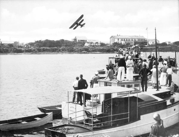 Slightly elevated view of a crowd of people standing on boats lined up along a pier to watch a bi-winged airplane flying over the water. Large buildings are on the opposite shoreline.