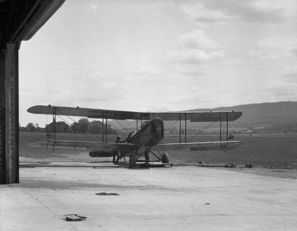 View from inside doorway of a hangar toward a man near an airplane at the landing field of an aerial mail station. Hills or mountains are in the distance.